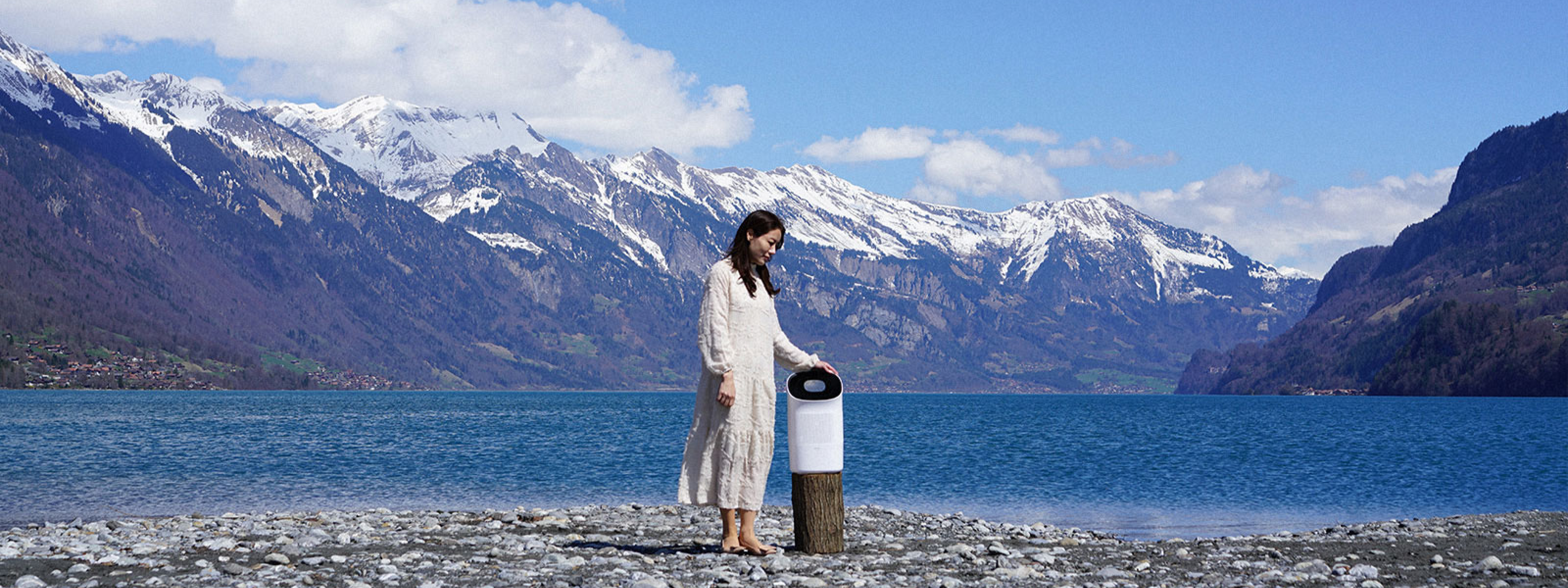 Woman standing on pebble beach next to Zenwell Super Air Purifier + Humidifier in front of alpine lake